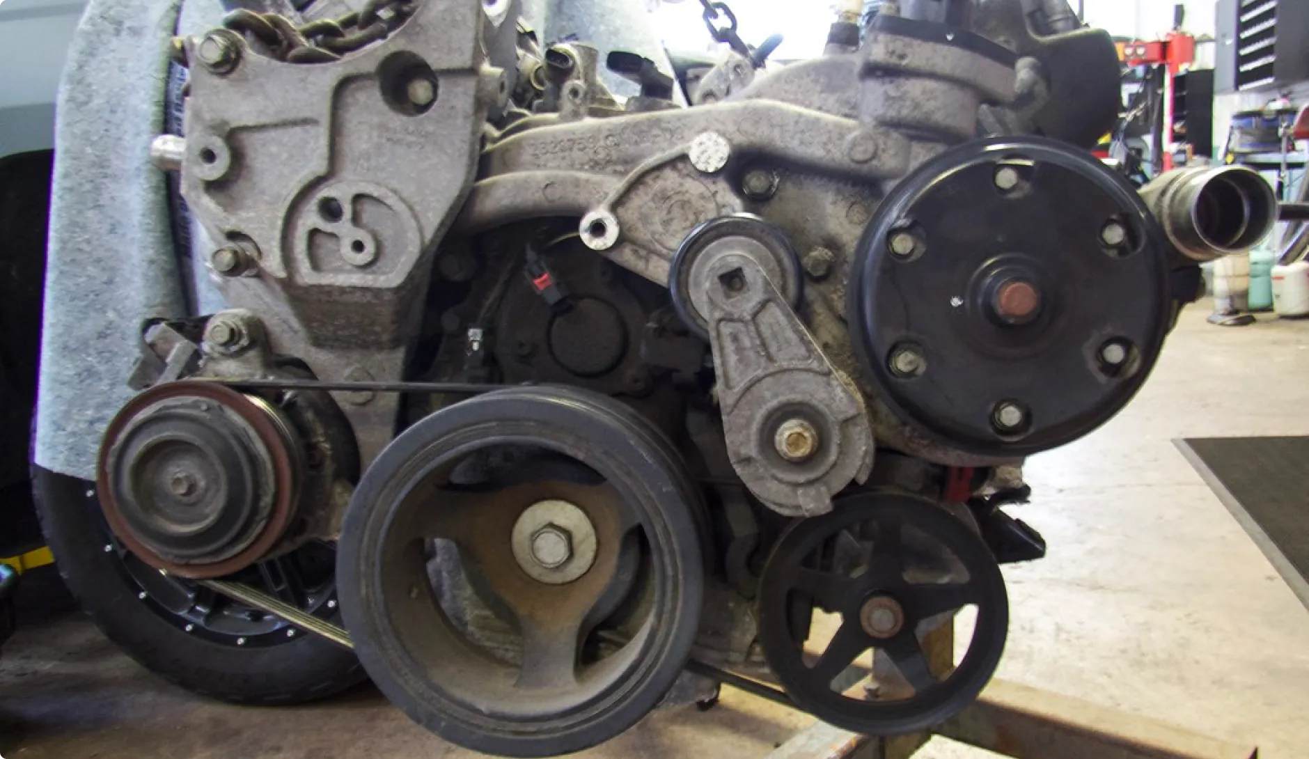Allied Automotive: Close-up of a car engine with visible belts, pulleys, and various components. The engine is mounted on a stand in a workshop with a concrete floor and blurred tools in the background.