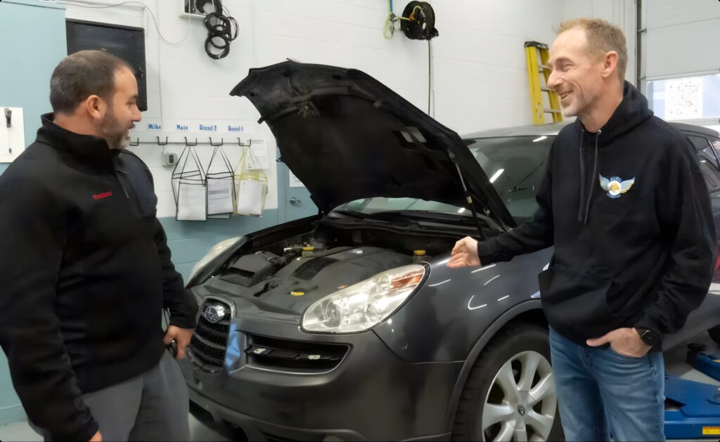 Allied Automotive: Two men are standing in a garage beside a car with its hood open, engaged in conversation. One man is gesturing towards the car while both appear to be smiling. Various tools and documents are visible on the walls behind them.