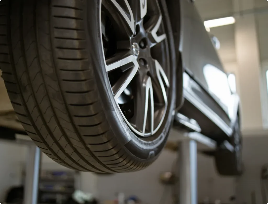 Allied Automotive: A close-up view of a car tire with alloy wheels, elevated on a hydraulic lift in a well-lit auto repair shop. The focus is on the tire tread and the underside of the car can be slightly seen.