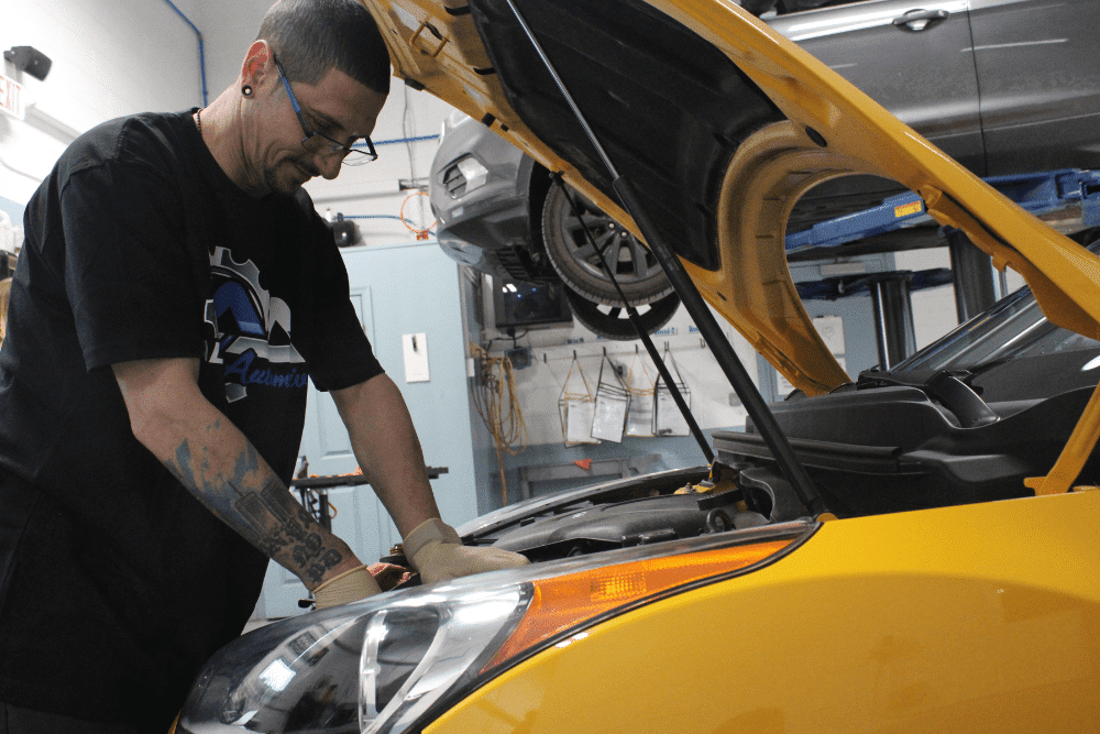 Auto mechanic working under the hood of a yellow vehicle at Allied Automotive in Allentown, PA, performing diagnostics or summer car maintenance service in a professional repair bay.